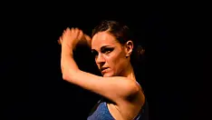 Ballerina di danza moderna in posa espressiva su sfondo scuro. Indossa un top blu senza maniche e una gonna fluida coordinata, con le braccia sollevate e lo sguardo intenso. L'illuminazione drammatica mette in risalto il movimento e la forza emotiva della performance